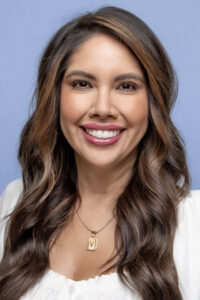A smiling woman with long, wavy brown hair, wearing a white top and a necklace, against a light blue background.